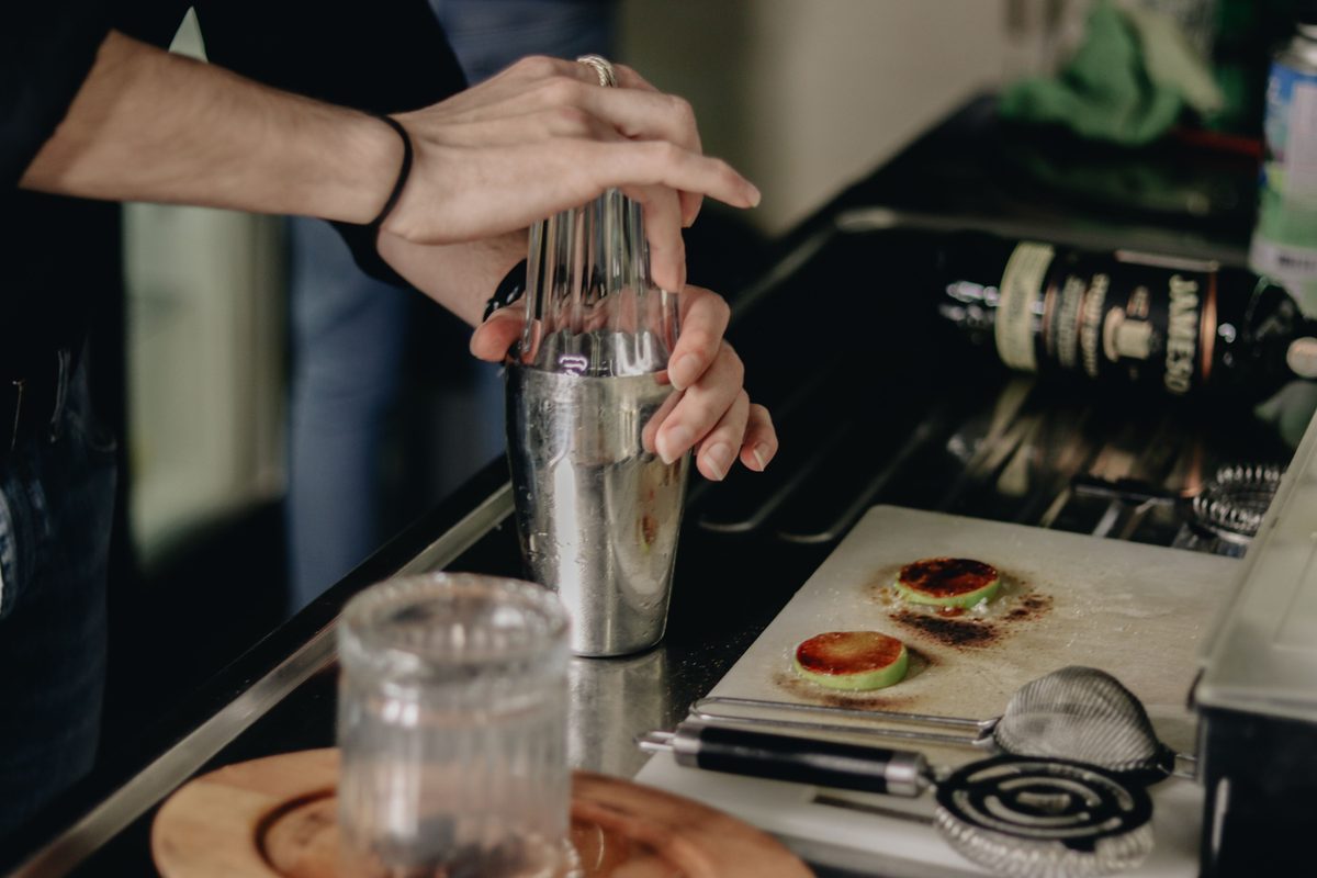 Hands straining a cocktail from a shaker at a bar laid with garnishes