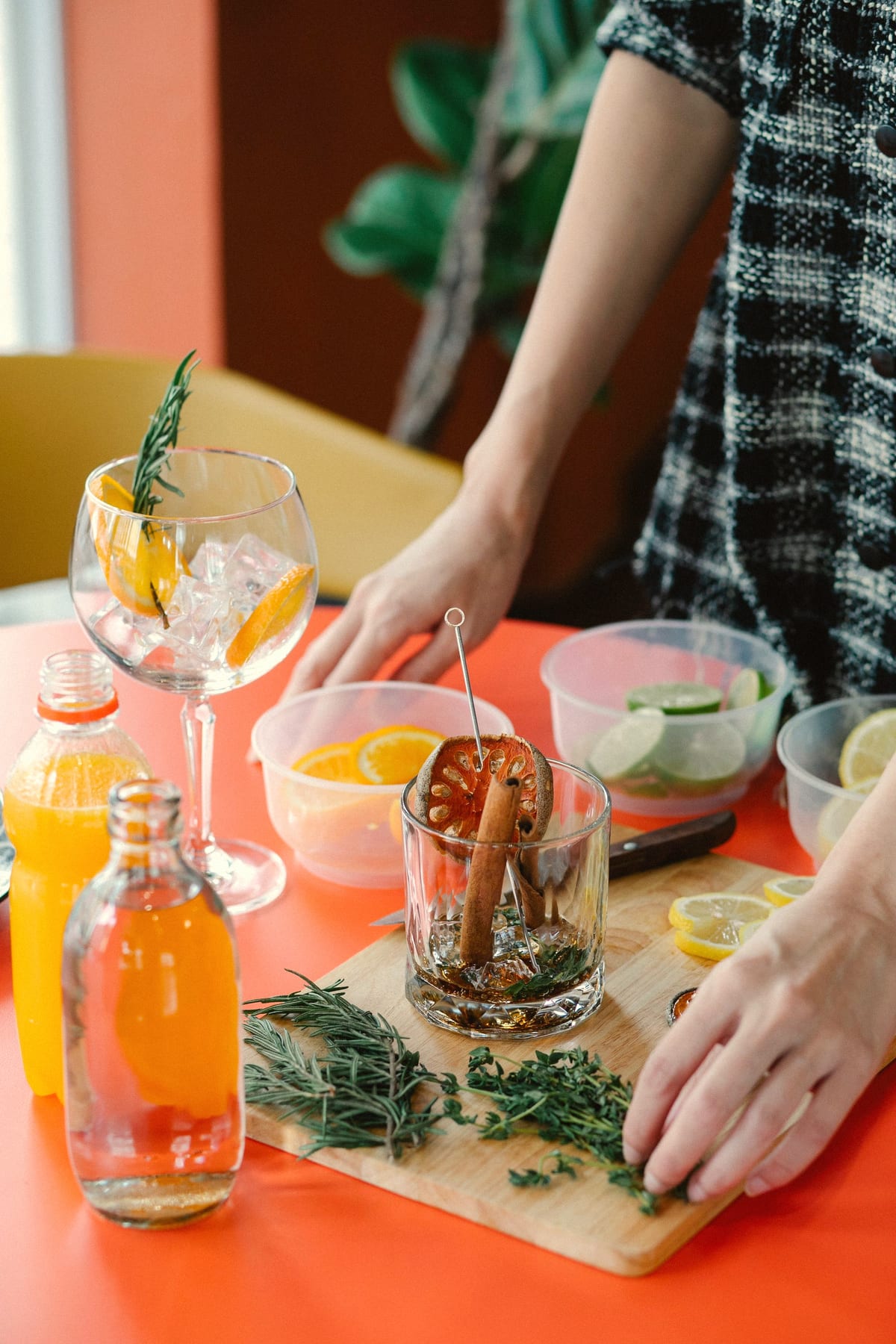 Hands arranging rosemary, thyme and citrus slices beside cocktail glasses on an orange table