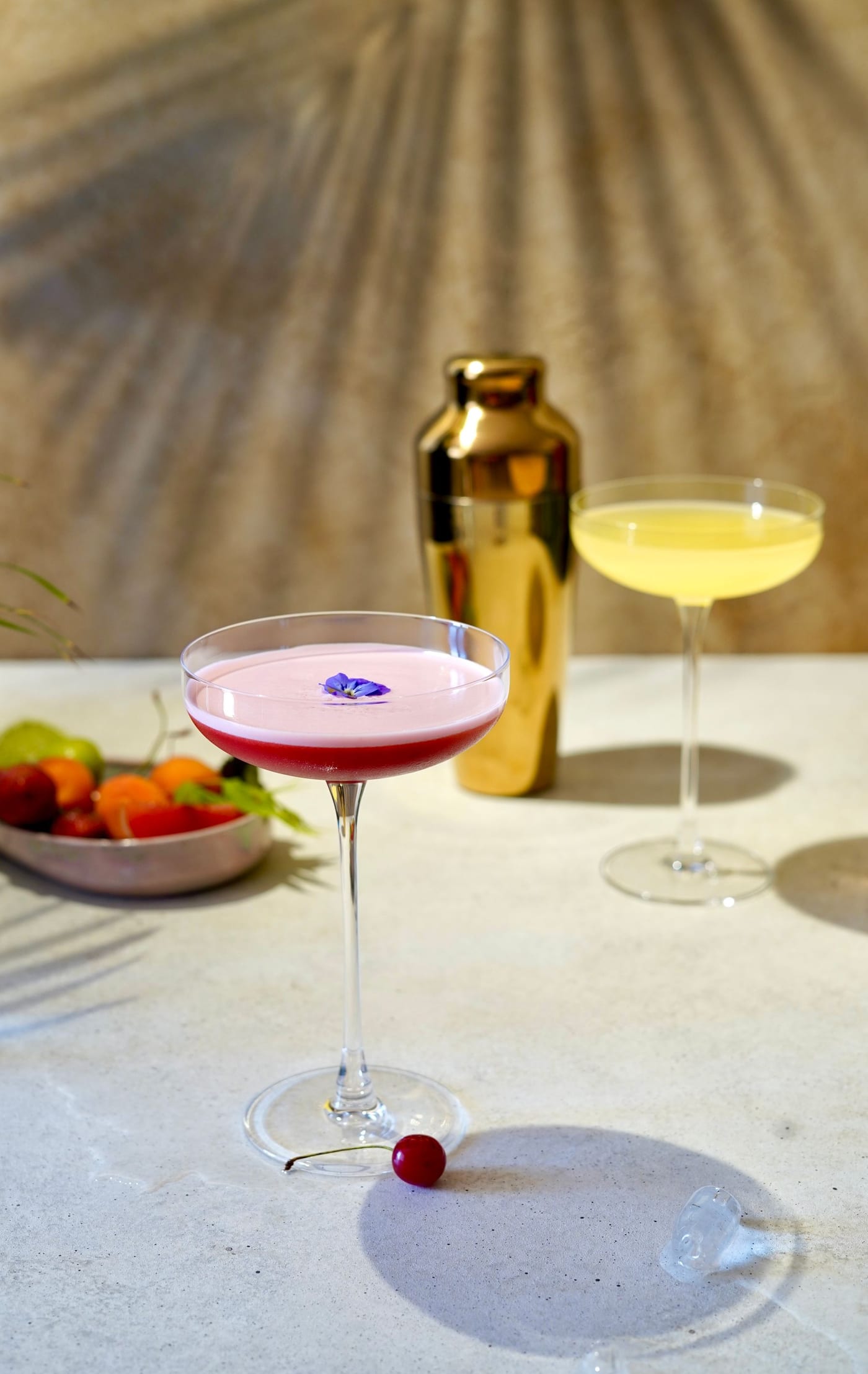A styled pink sour and a pale cocktail in coupes beside a gold shaker and fresh fruit