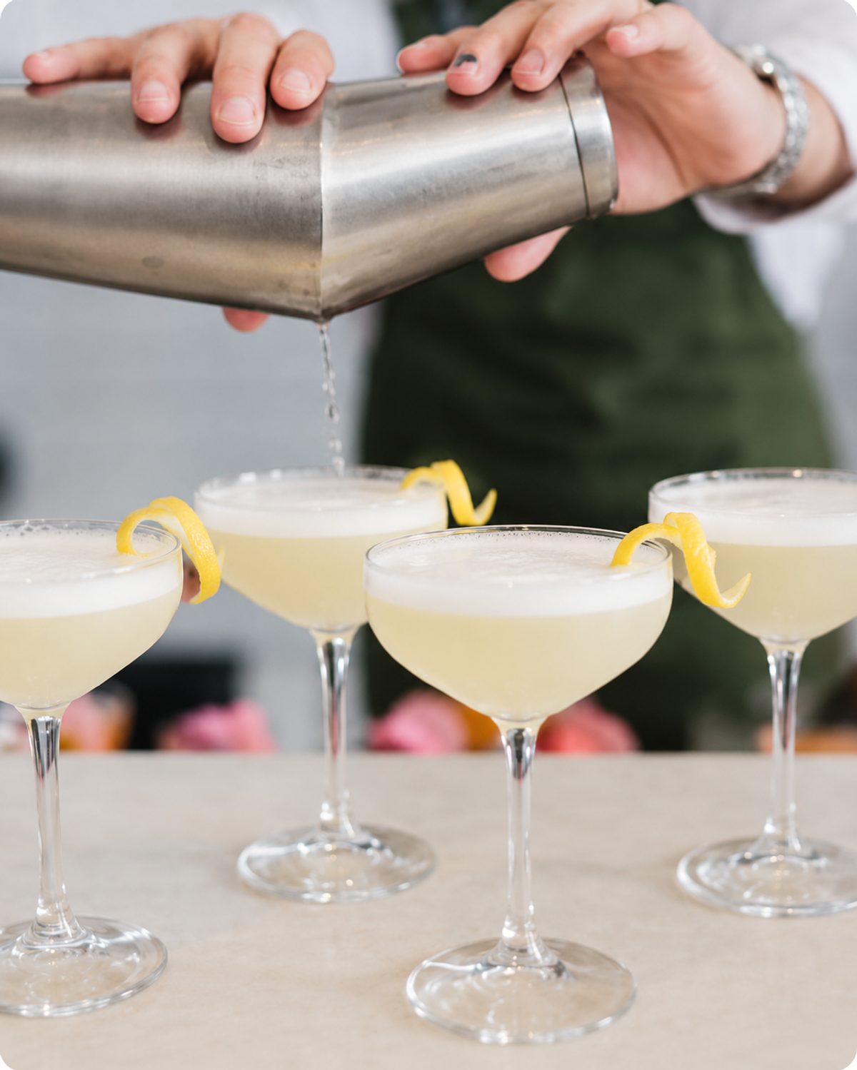 A cocktail shaker pouring a pale sour into four coupes garnished with lemon twists