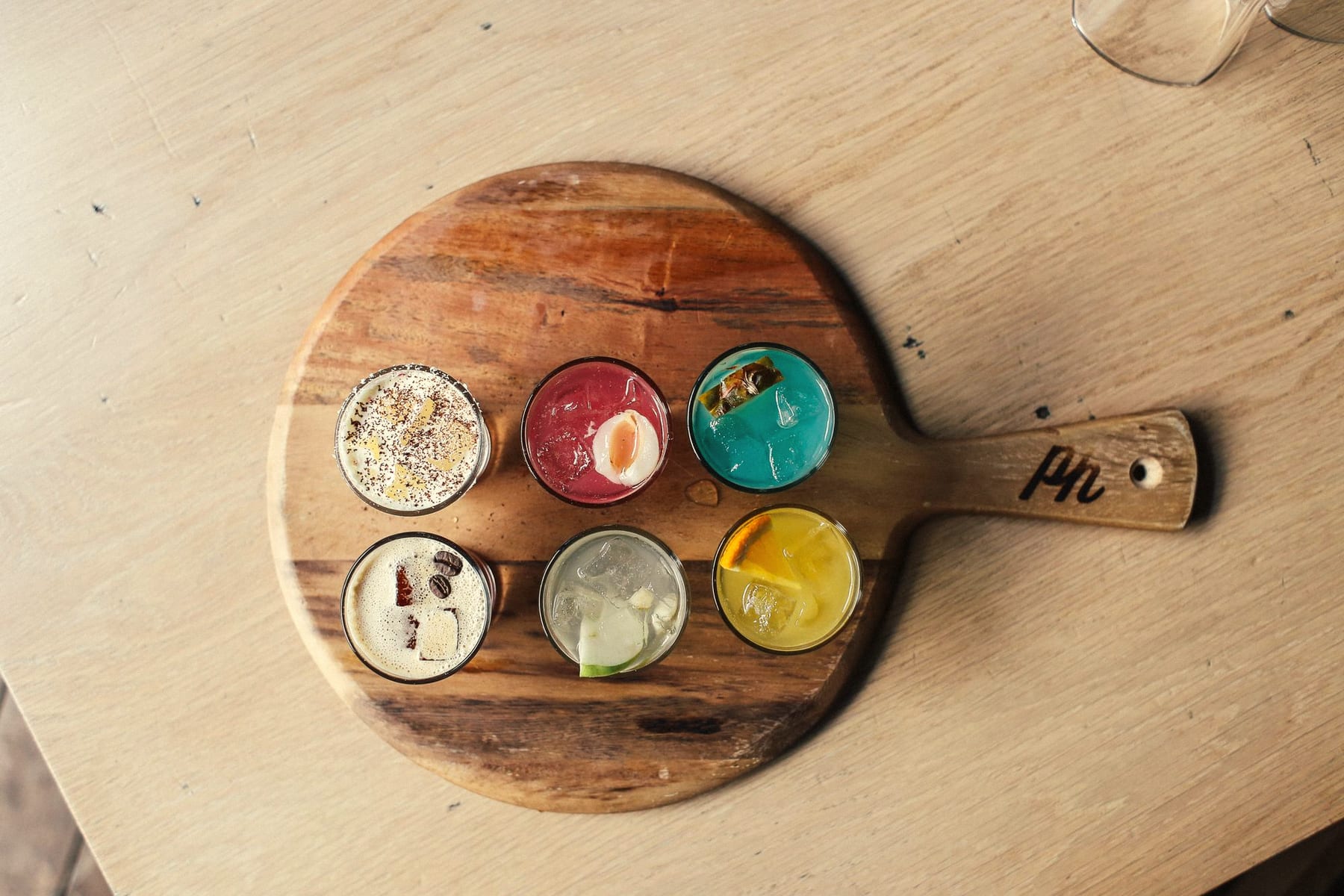 Overhead view of six colourful cocktails in small glasses on a round wooden board