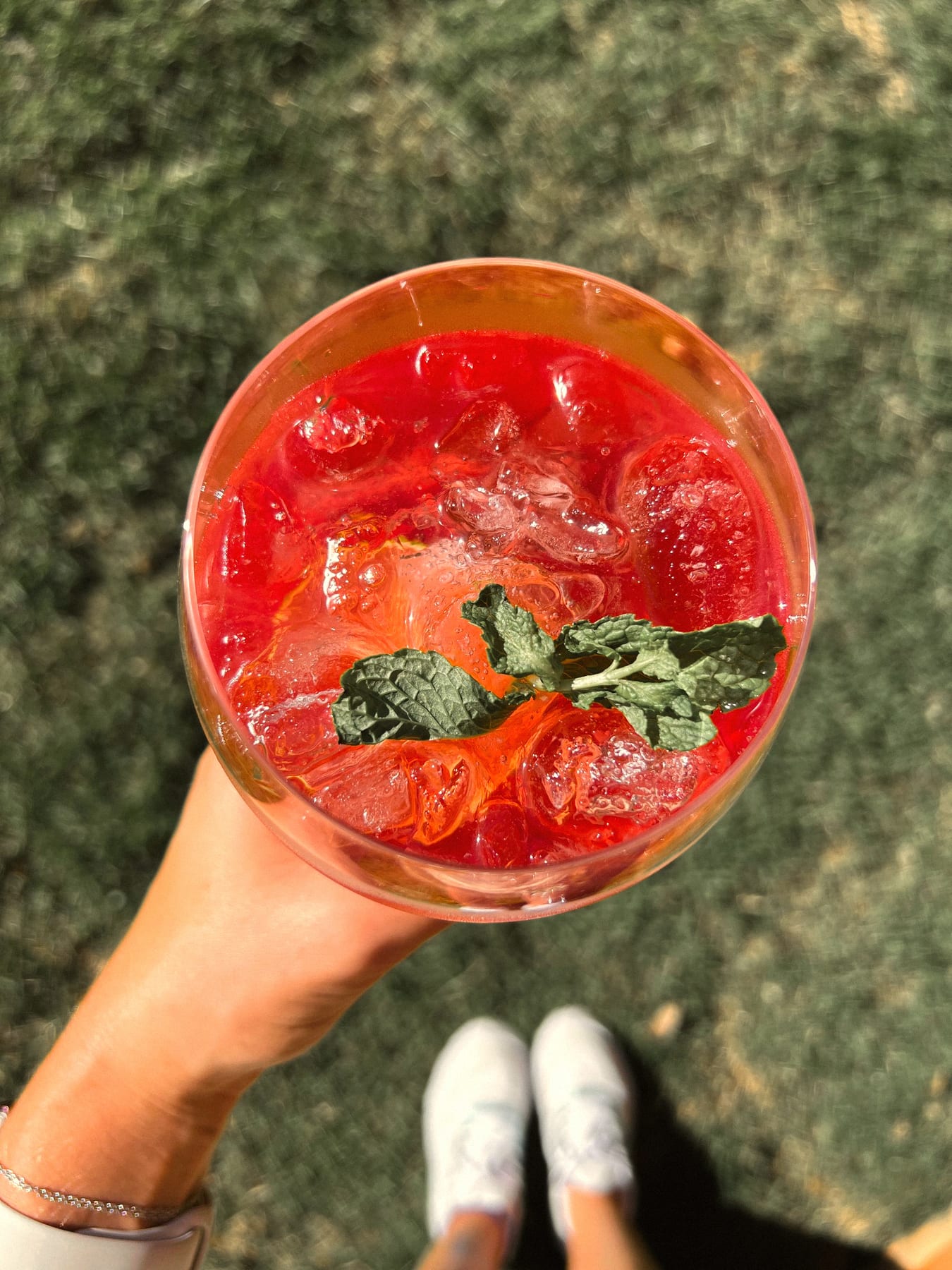 Hand holding a red cocktail with ice and mint above grass, white trainers below