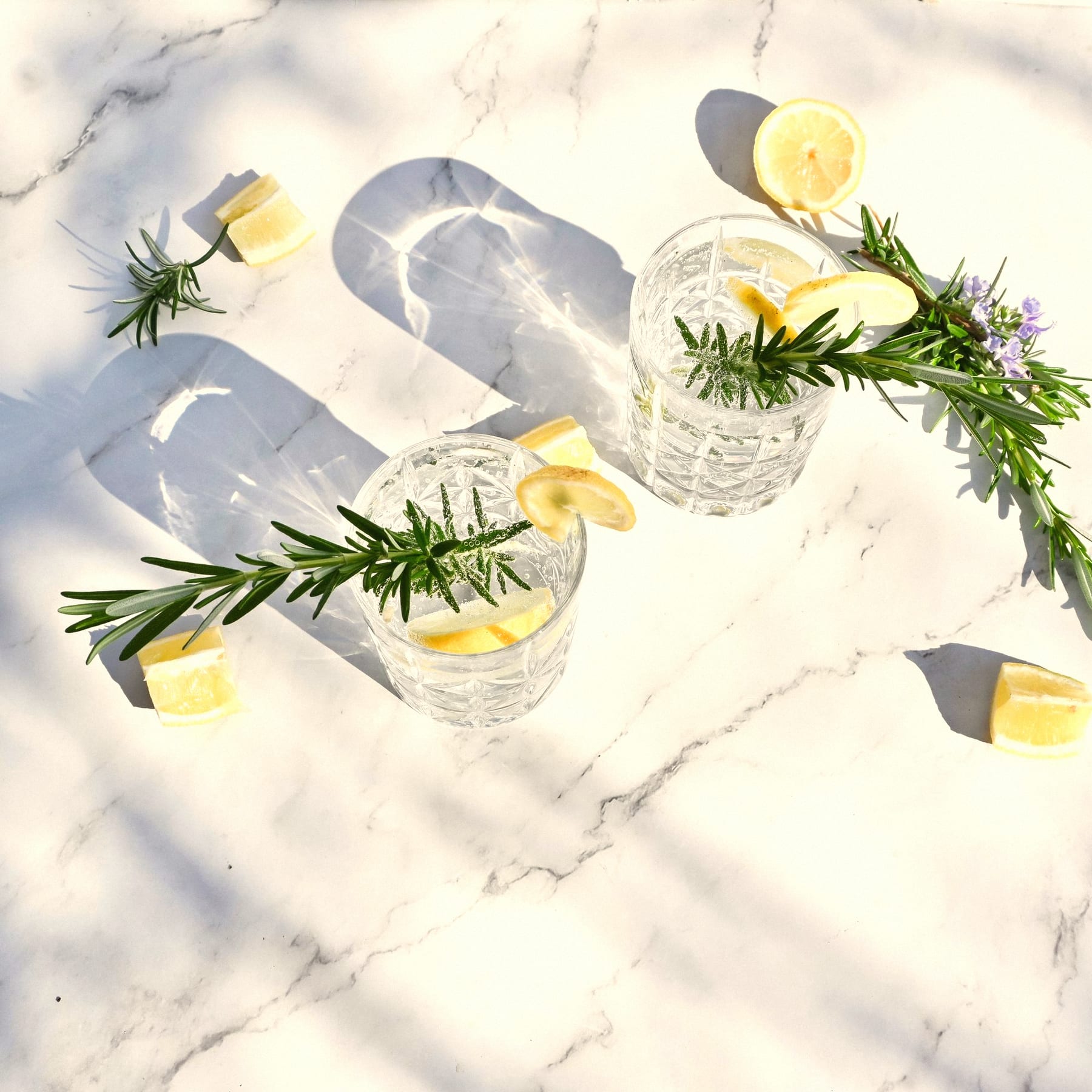 Two gin and tonics in cut-glass tumblers with lemon and rosemary on sunlit marble