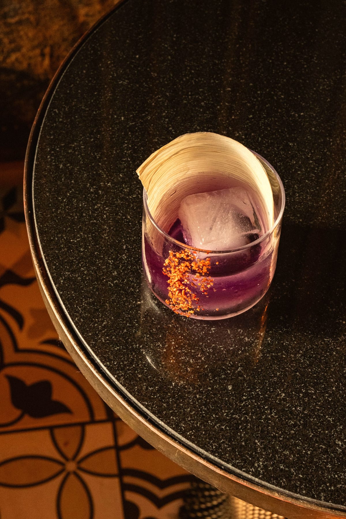 Purple drink with a large ice cube, red salt and a curled husk on a granite table