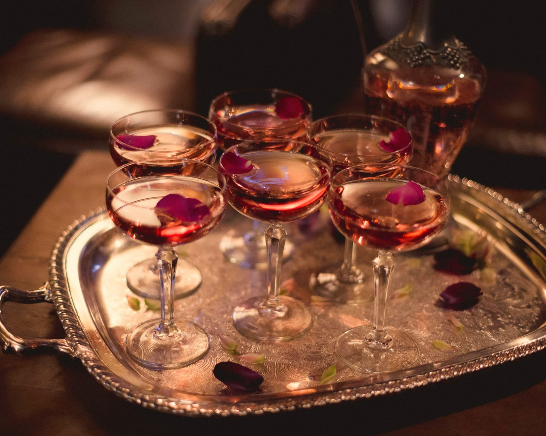 Six rosé coupes topped with rose petals on an ornate silver tray beside a decanter