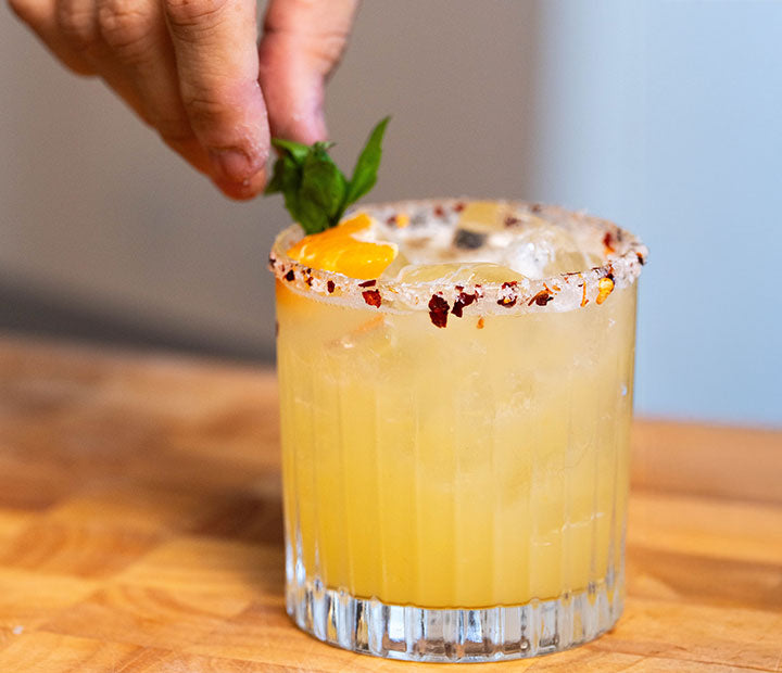 A hand placing a basil leaf on a hot honey margarita with a chilli-flake salt rim and an orange slice