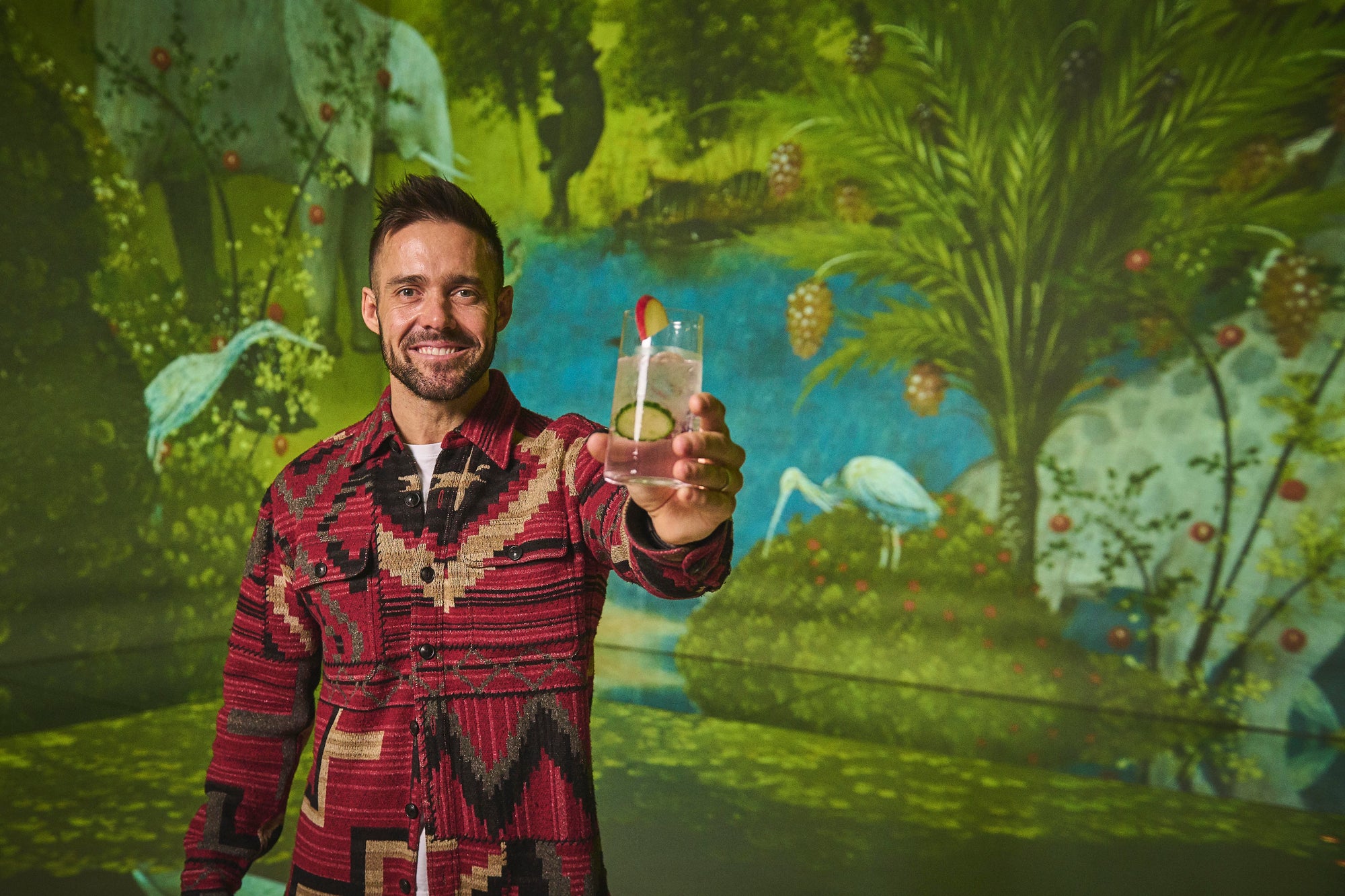Spencer Matthews holding a garnished clear cocktail in front of a projected jungle painting at Frameless