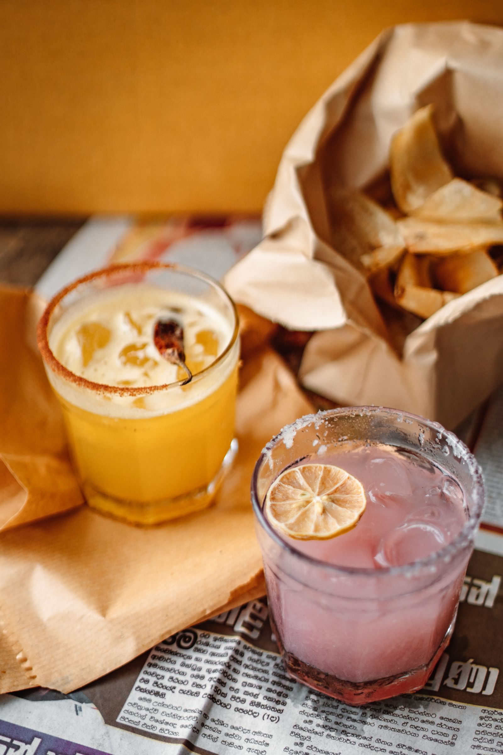 A chilli-rimmed yellow cocktail and a pink cocktail with a dried lime, in rocks glasses beside a paper bag of chips