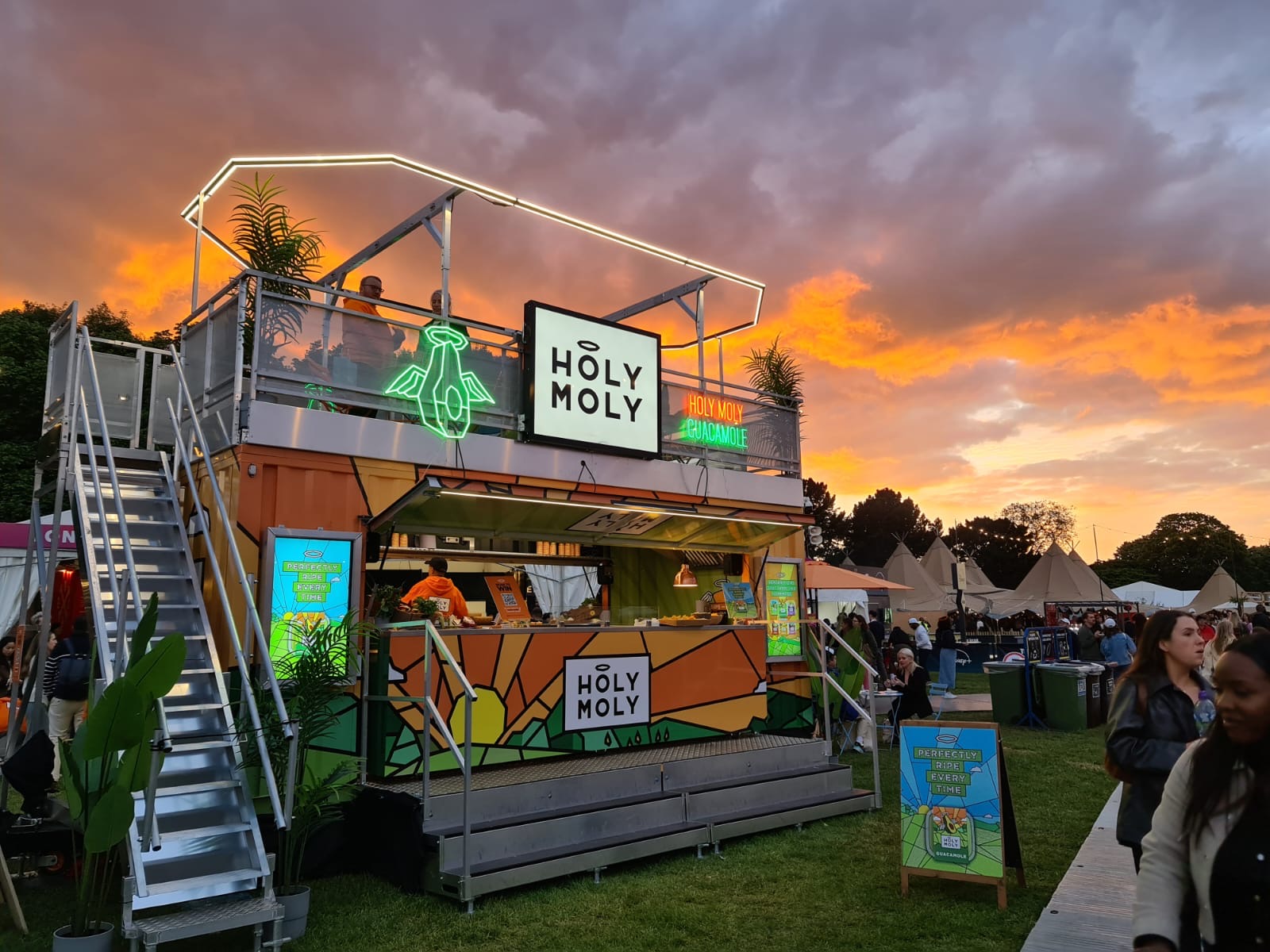 The Holy Moly guacamole stand at Taste of London: a two-storey orange container with neon signs and a rooftop deck under a sunset sky