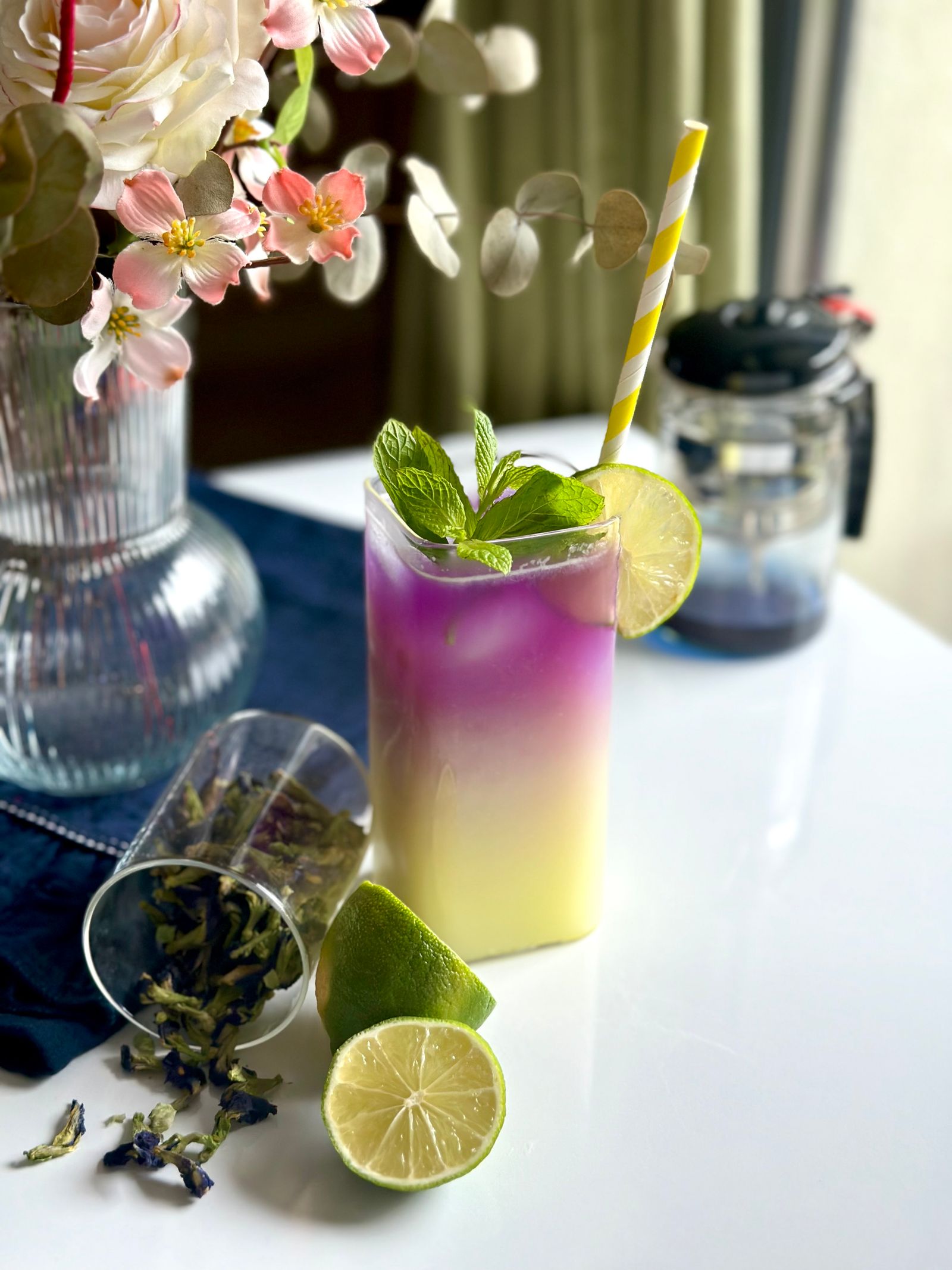 A tall glass of mint-topped drink shading from purple to pale yellow, with limes and dried butterfly pea flowers