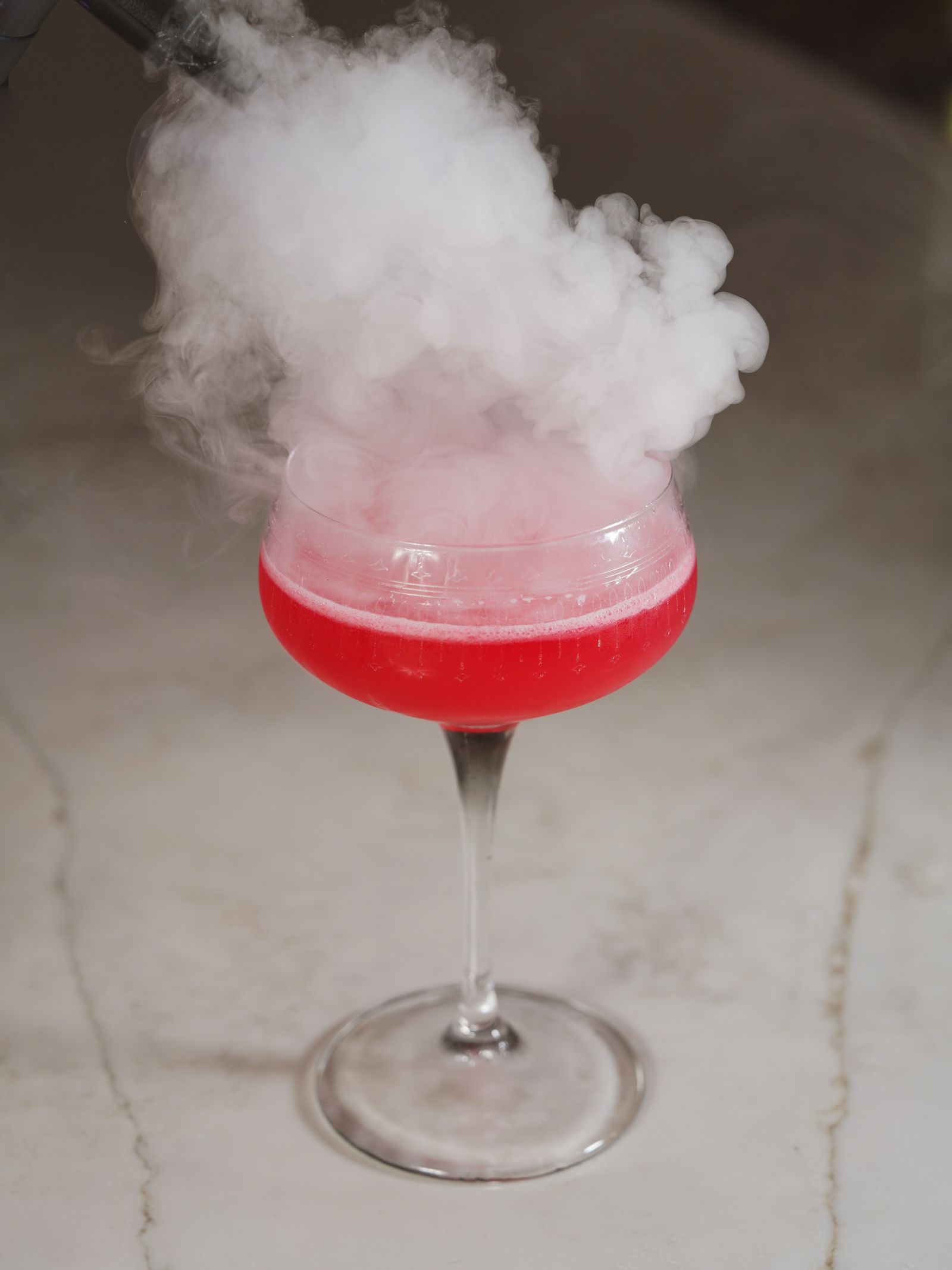A smoking red cocktail in a coupe glass on a marble table