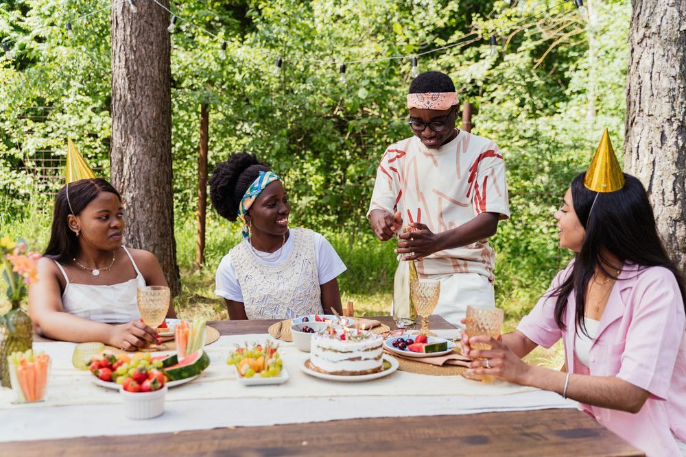 Friends at an outdoor party table with a cake and drinks among the trees