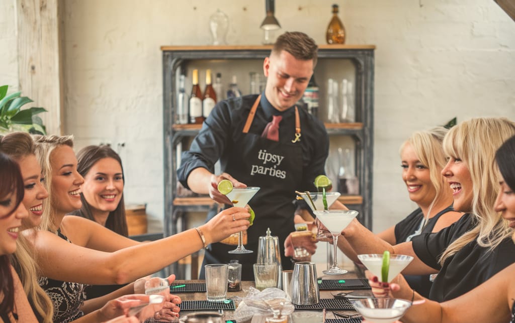 Bartender in a tipsy parties apron serving margaritas to a smiling group raising glasses