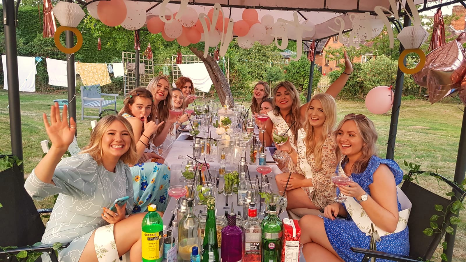 Hen party waving and raising pink cocktails at a long table under a decorated gazebo