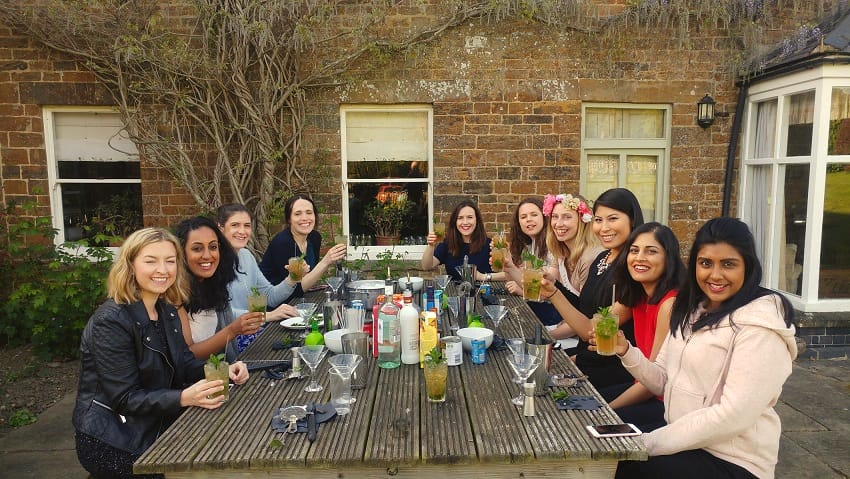 Group at an outdoor wooden table raising mint-garnished cocktails in front of a brick house