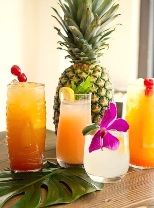 Tiki-glass cocktails with cherries and an orchid-topped drink in front of a whole pineapple
