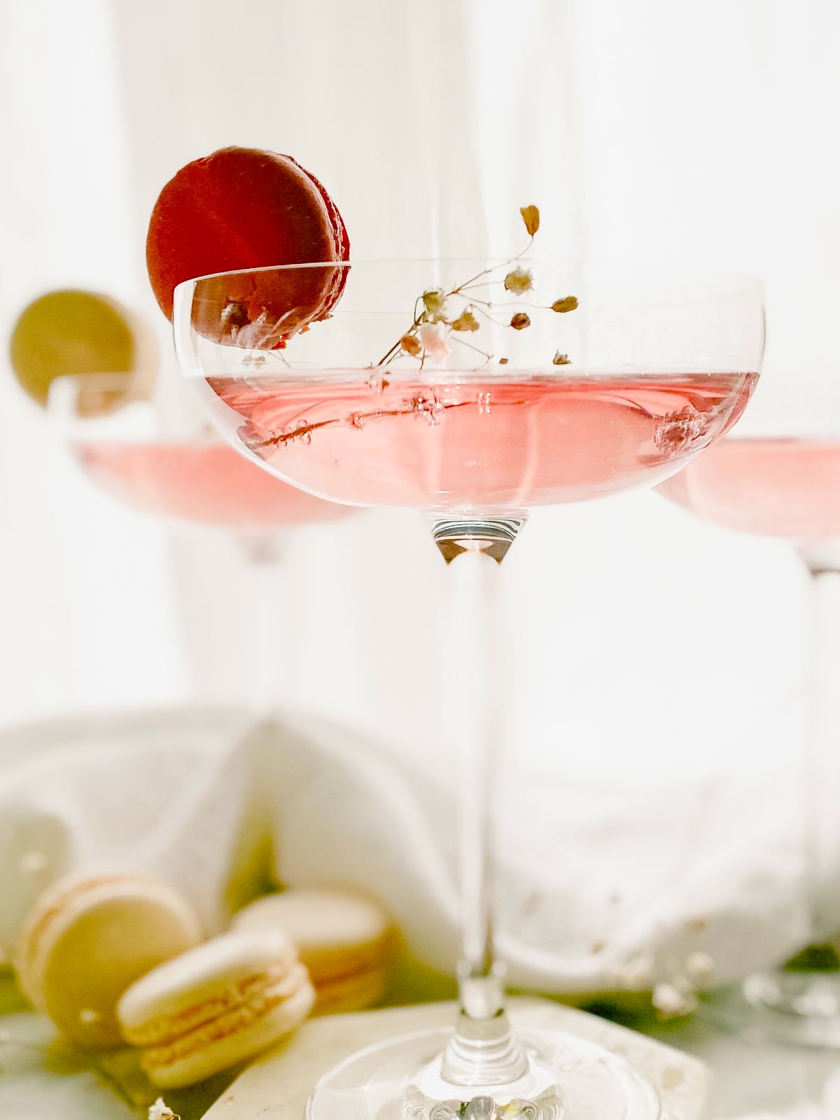 A pink fizz cocktail in a coupe with flowers and macarons