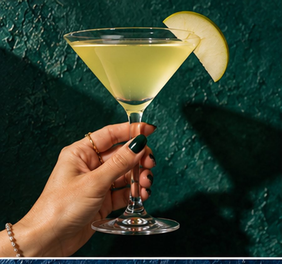 A hand holding an appletini with a green apple slice against a dark green wall
