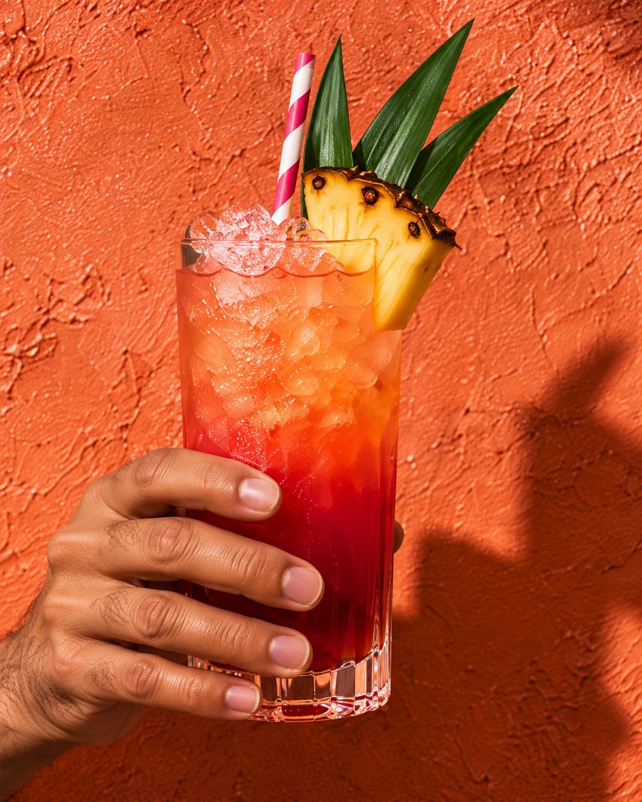 A hand holding a red and orange highball with pineapple leaves and a striped straw against an orange wall