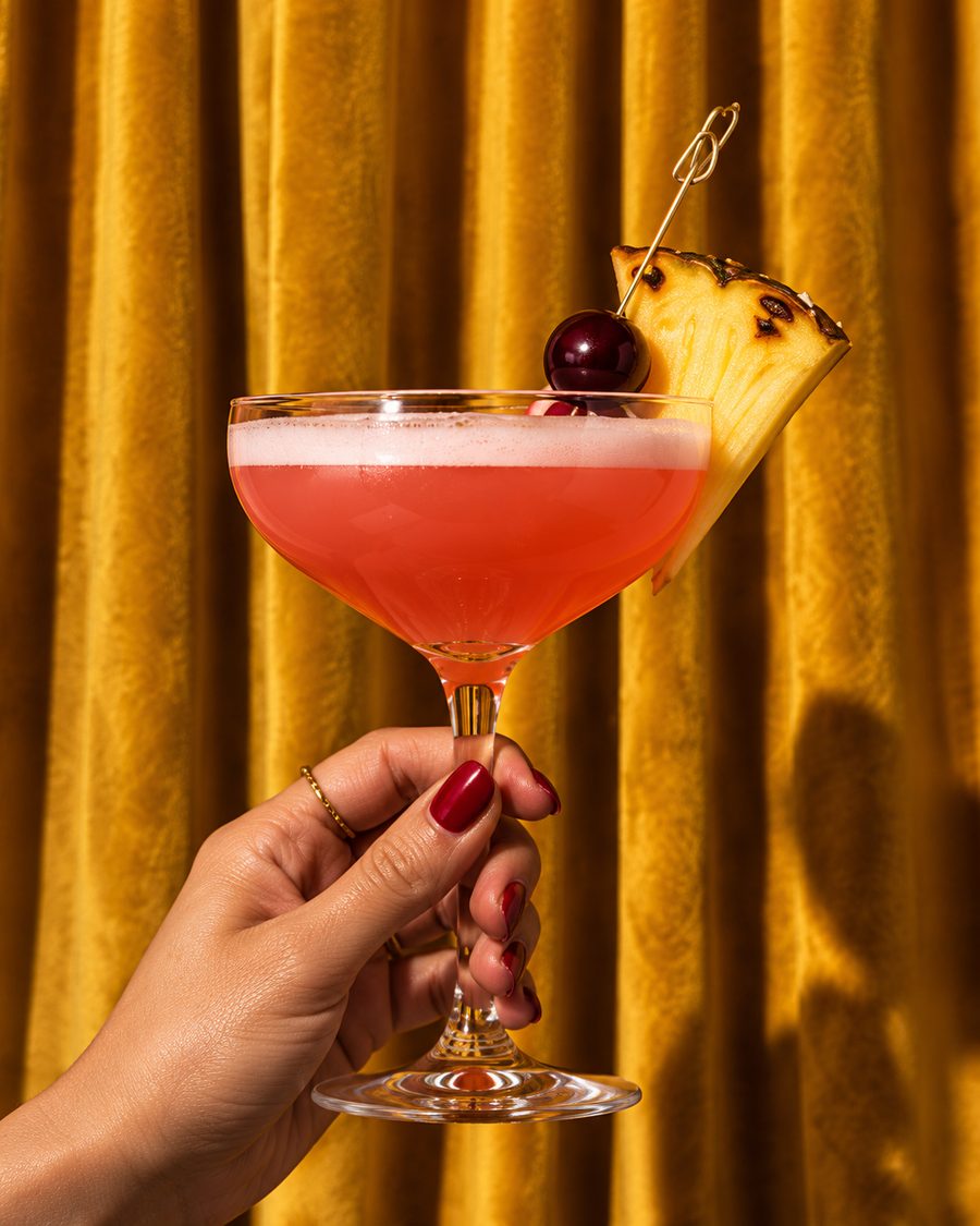 A hand holding a pink sour with a pineapple wedge and cherry against a gold curtain