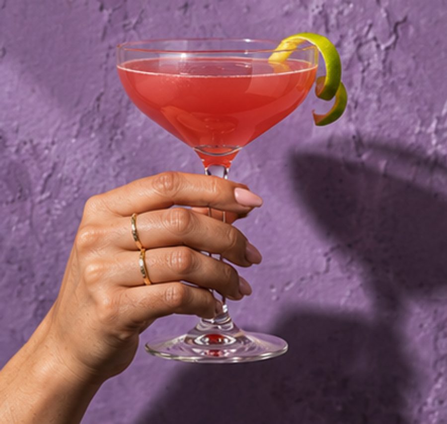 A hand holding a cosmopolitan with a lime twist against a lilac wall