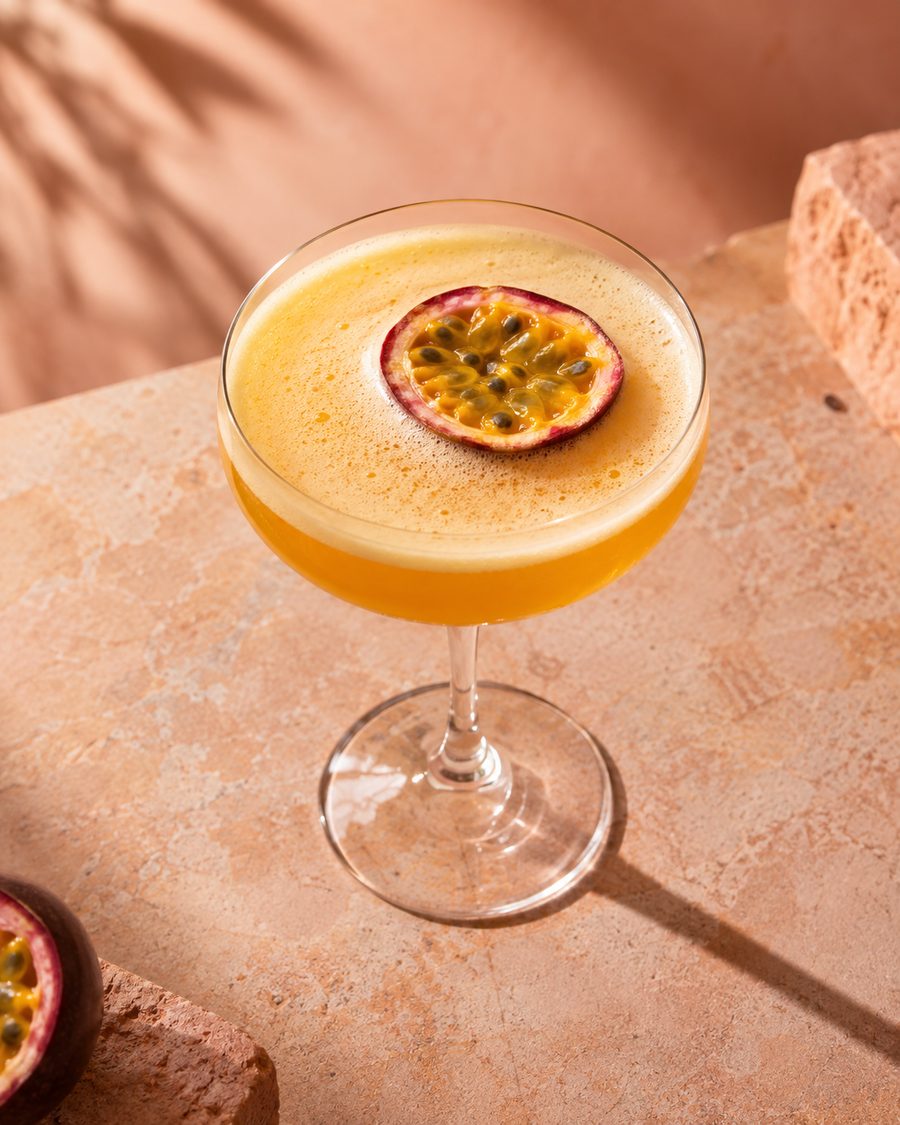 A passion fruit martini on a peach surface in dappled sun