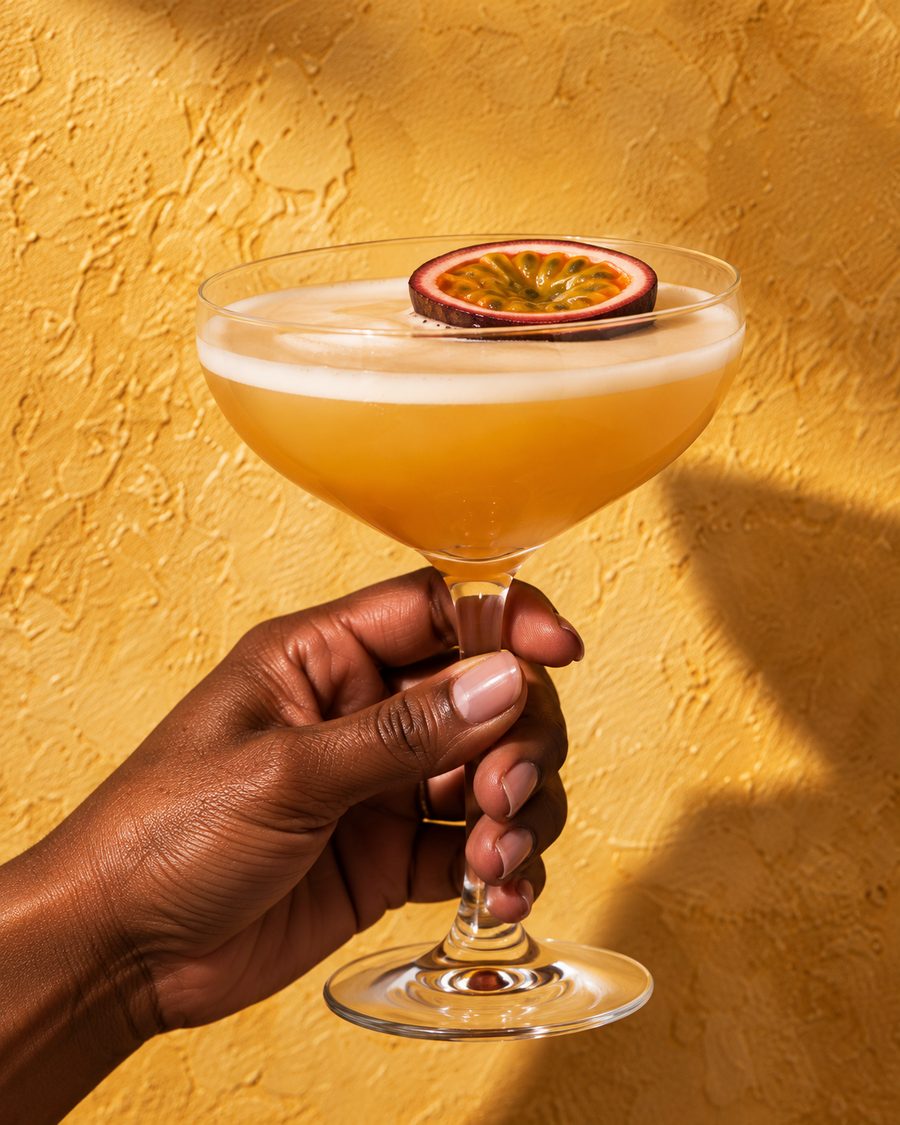 A hand holding a passion fruit martini by the stem against a mustard yellow wall