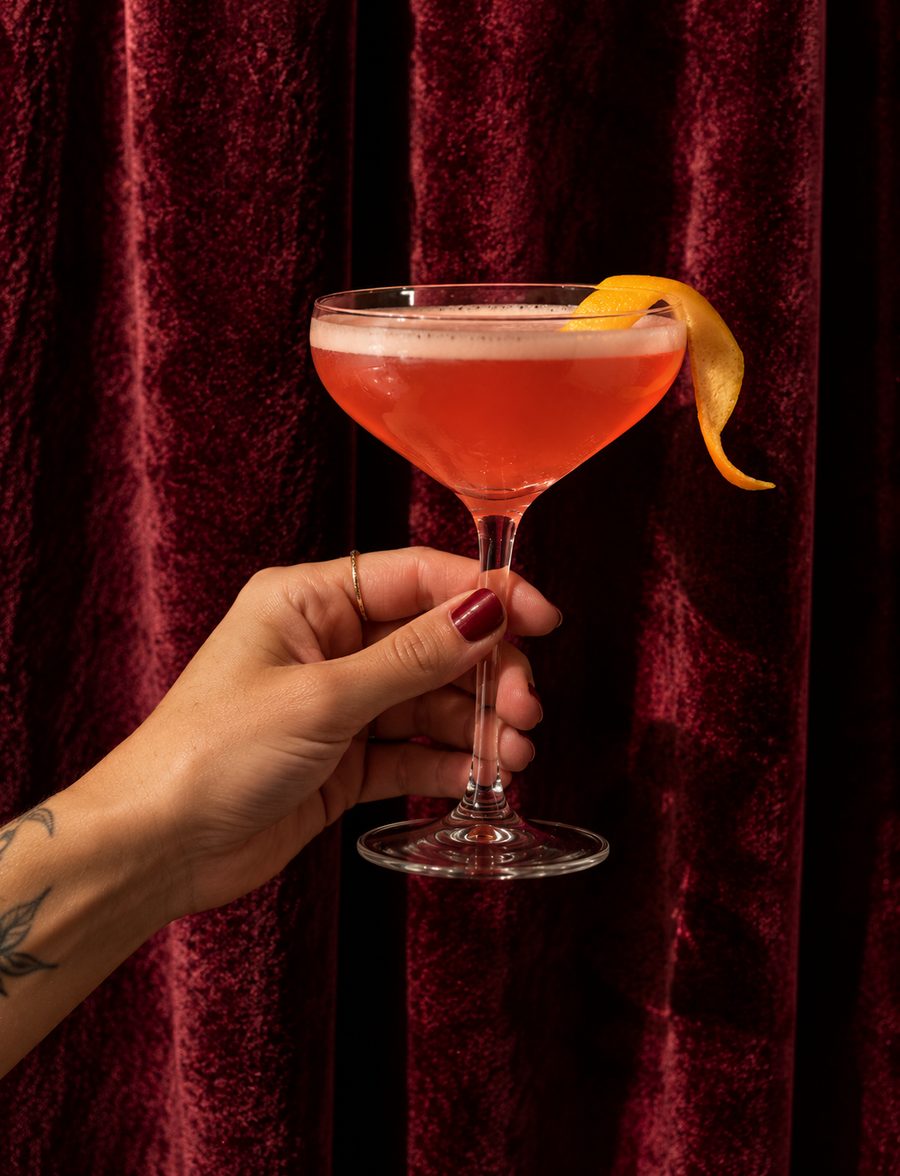 A hand holding a coral sour in a coupe by the stem, with an orange twist, against red velvet