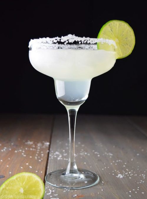 A Margarita cocktail garnished with a lime wheel served in a Margarita glass on a wooden floor with a lime also on the floor.