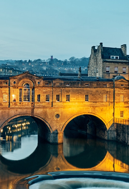 Night time view of the iconic Putney Bridge in Bath, with warm up-lights lighting the stone structure, reflecting beautifully on the river below